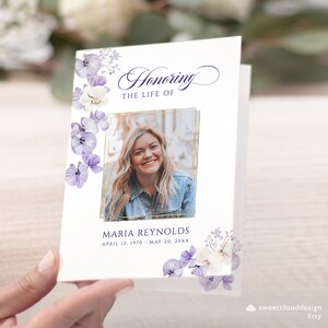 Op de afbeelding: Een witte herdenkingskaart met een gouden lijst om een foto van een vrouw. De kaart is versierd met paarse en witte bloemen aan de linker- en rechterkant. De tekst "Honoring the Life of Maria Reynolds April 12, 1970 - May 20, 20XX" staat op de kaart gedrukt.
