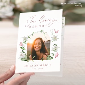 Op de afbeelding: Een witte rouwkaart met een rand van een bloemkrans en een foto van een lachende vrouw. De tekst "In loving memory" is in roze schrift geschreven. De naam "Emilia Anderson" en de data "1995 - 2028" staan onder de foto.
