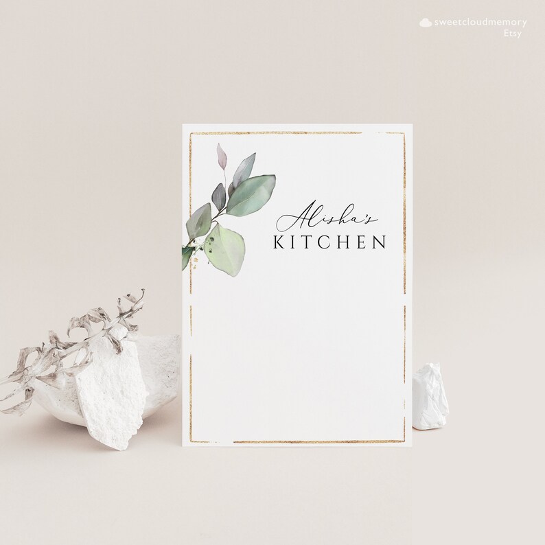 Op de afbeelding: Een witte kaart met een gouden rand en een groen bladerenontwerp. De tekst "Alisha's Kitchen" is in zwart gedrukt.