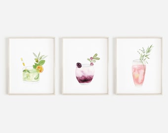 May include: Three watercolor paintings of cocktails in frames. The first painting shows a green cocktail with a lime wedge and a yellow straw. The second painting shows a purple cocktail with berries and a blackberry. The third painting shows a pink cocktail with a sprig of greenery.