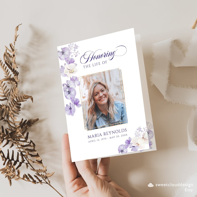 Op de afbeelding: Een witte rouwkaart met een gouden lijst en een foto van een vrouw. De kaart is versierd met paarse en witte bloemen en de tekst "Honoring the Life of Maria Reynolds April 12, 1970 - May 20, 20XX"