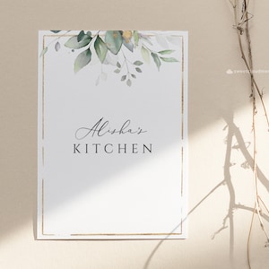 Op de afbeelding: Een wit bedrukbaar bord met een gouden lijst en groene aquarelbladeren. De tekst op het bord luidt "Alisha's Kitchen".