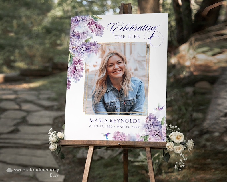 May include: A white memorial sign with a purple floral design and a photo of a woman. The sign reads "Celebrating the Life" and "Maria Reynolds, April 12, 1980 - May 20, 20XX".