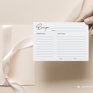 May include: A white recipe card with the word "Recipe" written in cursive at the top. The card has lines for writing down ingredients, directions, servings, and time. The card is held in a hand against a light pink background.
