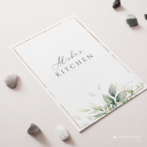 Op de afbeelding: Een witte kaart met een gouden rand en een groen aquarel bloemenontwerp. De tekst "Alisha's Kitchen" is geschreven in zwarte cursieve letters.