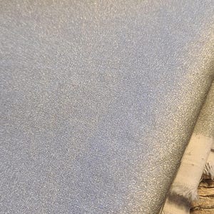 May include: A folded piece of silver fabric with a metallic, shimmering finish. The material features a fine, woven texture and visible frayed edges along the side, showcasing the light-reflecting quality of the silver-toned surface throughout.