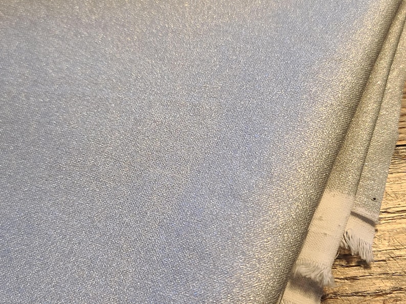 May include: A folded piece of silver fabric with a metallic, shimmering finish. The material features a fine, woven texture and visible frayed edges along the side, showcasing the light-reflecting quality of the silver-toned surface throughout.
