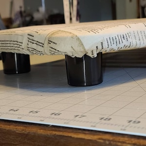 May include: An ironing board with a cream-colored cover printed with newspaper text. The board is supported by four short, glossy black legs. The board is positioned on a white cutting mat with a grid pattern.