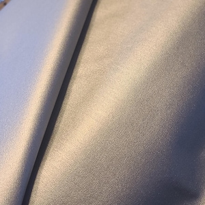 May include: A close-up of a smooth, light gray fabric with a subtle sheen. The fabric is folded in half, creating a diagonal line across the image.
