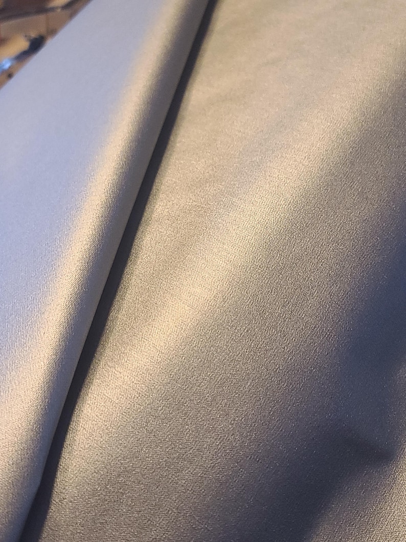 May include: A solid light gray fabric with a smooth, matte texture. The material is folded over, creating a diagonal crease that highlights the consistent color and fine weave of the textile surface, showing a soft sheen under the light.
