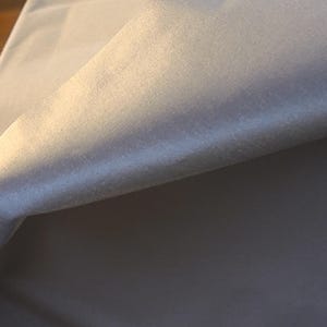 May include: A piece of solid white fabric with a smooth, slightly reflective surface texture. The material is folded over, creating a soft curve and a gradient of light and shadow across the surface, highlighting the clean, plain white finish.