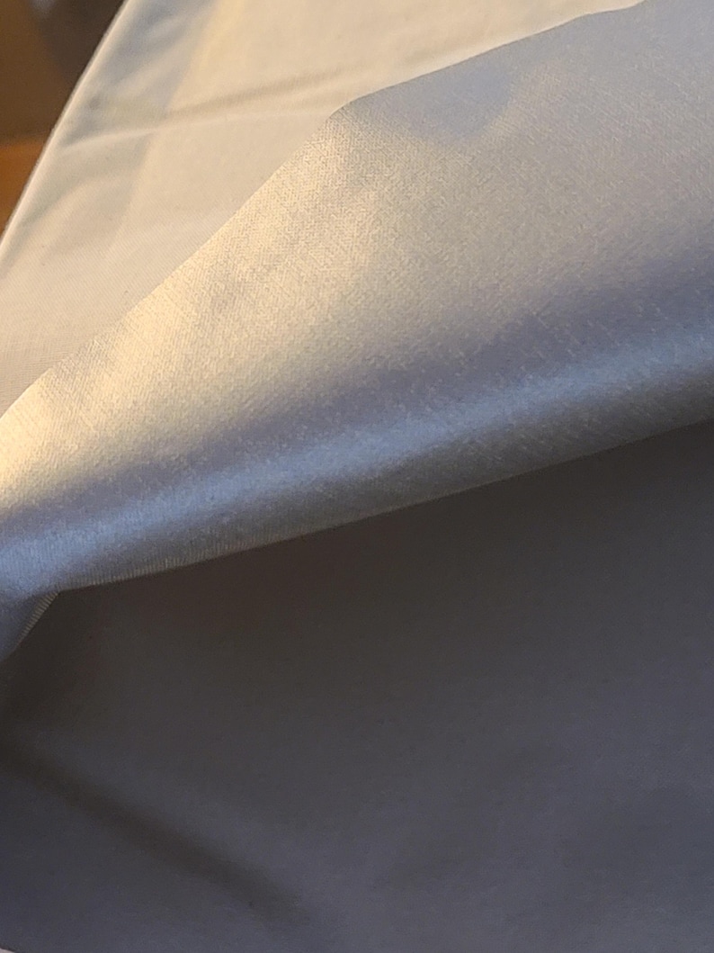 May include: A piece of solid white fabric with a smooth, slightly reflective surface texture. The material is folded over, creating a soft curve and a gradient of light and shadow across the surface, highlighting the clean, plain white finish.