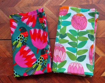 Könnte beinhalten: Zwei Stoffservietten mit tropischem Blumen- und Vogeldesign. Eine Serviette ist grün mit rosa Blüten und schwarzen und grünen Vögeln. Die andere Serviette ist weiß mit rosa Blüten und grünen Blättern.