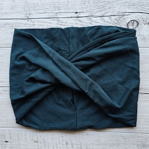 Pode incluir: Uma faixa de cabelo estilo turbante em verde-azulado escuro. O tecido é macio e parece ser malha de jersey. A faixa é torcida na frente, criando um acessório elegante e confortável. O fundo é uma superfície de madeira clara.