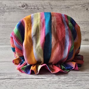 Puede incluir: Un gorro a rayas de colores en tonos rojos, naranjas, amarillos, verdes, azules y morados. La tela tiene rayas verticales y un borde con volantes. El gorro está hecho de un material suave y ligero.