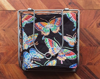 May include: A silver metal napkin holder with a black fabric liner. The fabric has a colorful butterfly pattern with red, blue, yellow, and green accents.