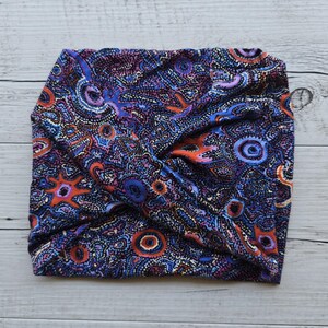 Op de afbeelding: Een blauw, oranje en roze gestreept stoffen hoofdband met een gedraaid ontwerp. De stof heeft een druk patroon van stippen, cirkels en swirls.