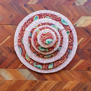 May include: A set of five nested, circular fabric placemats. The largest is a light pink with a white floral pattern. The others have a burgundy paisley design with teal and gold accents. The placemats are arranged on a wooden surface.