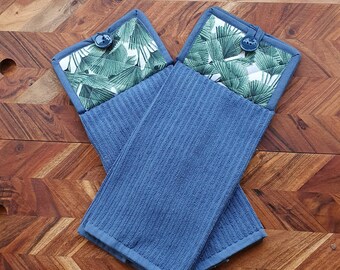 May include: Two blue kitchen pot holders with a green and white tropical leaf pattern. The pot holders have a button on the top.