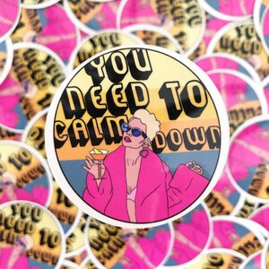 May include: A round sticker with a yellow background and the text "You Need To Calm Down" in black. A cartoon illustration of a woman with blonde hair wearing a pink robe and sunglasses is holding a martini glass.