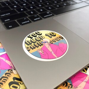 May include: A round sticker with a yellow background and the text "You Need To Calm Down" in black and white letters. A cartoon illustration of a woman with blonde hair wearing a pink robe and sunglasses is holding a glass of champagne.