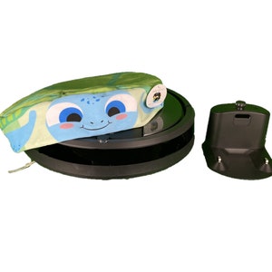 Puede incluir: Un robot aspirador negro con una cubierta de tela en forma de tortuga verde y azul. El robot aspirador está sobre una superficie blanca y tiene una base de carga a la derecha.