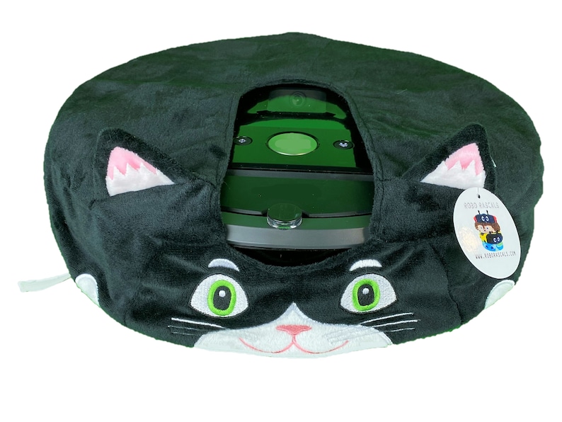 May include: Black and white plush cat-shaped cover for a robot vacuum cleaner. The cover has green eyes and a pink nose. The vacuum cleaner is visible through the opening in the cover. The cover has a tag that says "Roro Raccoon" and "www.rororaccoon.com".