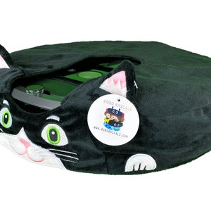 May include: Black plush cat-shaped cover for a round robot vacuum cleaner. The cover has white embroidered eyes, nose, and whiskers. The vacuum cleaner is partially visible underneath the cover. A white tag with the text "ROBO RASCALS" and a website address is attached to the cover.