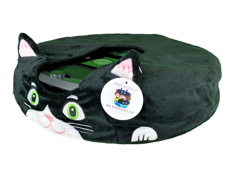May include: Black plush cat-shaped cover for a round robot vacuum cleaner. The cover has white embroidered eyes, nose, and whiskers. The vacuum cleaner is partially visible underneath the cover. A white tag with the text "ROBO RASCALS" and a website address is attached to the cover.