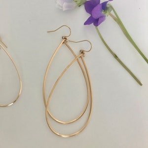May include: A pair of gold teardrop-shaped earrings with a double wire design. The earrings are hanging from a white surface.