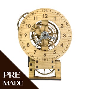 Peut inclure: Une horloge mécanique en bois brun clair avec un cadran circulaire affichant les chiffres de 1 à 24, des engrenages internes visibles et des aiguilles argentées. La structure repose sur une base rectangulaire en bois avec des engrenages.