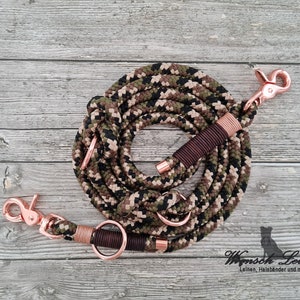 Könnte beinhalten: Eine Doppelleine für Hunde im Tarnmuster mit roségoldenen Beschlägen. Die Leine hat zwei Clips und zwei Ringe. Die Leine ist auf einer Holzoberfläche aufgerollt.