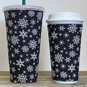 May include: Two reusable coffee cup sleeves with a black background and white snowflake pattern. The sleeve on the left is on a tall cup, while the sleeve on the right is on a shorter cup.