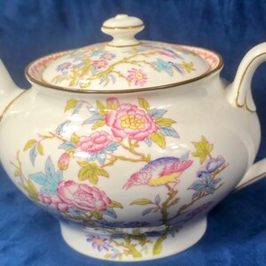 May include: A white ceramic teapot with a lid, decorated with a floral and bird pattern in pink, blue, and yellow. The teapot has a curved spout and handle, with gold trim. The design includes pink roses and colorful birds.