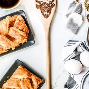 K&ouml;nnte beinhalten: Heller Holzl&ouml;ffel mit einem gravierten Design einer Highland-Kuh. Der L&ouml;ffel liegt auf einer wei&szlig;en Marmoroberfl&auml;che, umgeben von Geb&auml;ck, Tee und Eiern. Ein dekoratives K&uuml;chenutensil.