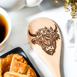 K&ouml;nnte beinhalten: Holzl&ouml;ffel mit einem lasergravierten Design einer Highland-Kuh, verziert mit floralen Akzenten. Der L&ouml;ffel ist hell und die Merkmale der Kuh sind detailliert. Teil eines K&uuml;chenutensilien-Sets.