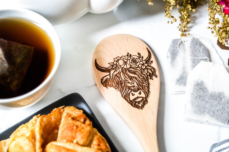 K&ouml;nnte beinhalten: Holzl&ouml;ffel mit einem lasergravierten Design einer Highland-Kuh, verziert mit floralen Akzenten. Der L&ouml;ffel ist hell und die Merkmale der Kuh sind detailliert. Teil eines K&uuml;chenutensilien-Sets.