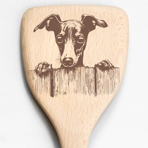 May include: A wooden spoon with a brown illustration of a dog peeking over a wooden fence. The dog has large ears and is looking directly at the viewer. The spoon is made of light-colored wood.