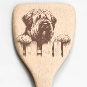 May include: Wooden kitchen spatula with a brown illustration of a dog peering over a wooden fence. The dog has long, shaggy fur and is looking directly at the viewer. The spatula is made of light-colored wood.