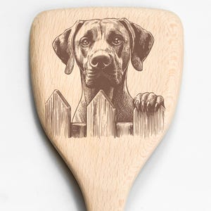 May include: Wooden spoon with a detailed engraving of a dog's face peering over a picket fence. The dog is brown, and the fence is a lighter shade of brown. The spoon is made of light-colored wood.