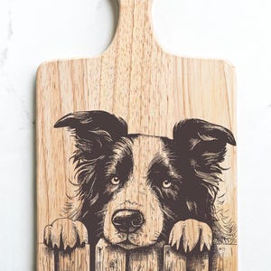 May include: Wooden cutting board with a handle, featuring a detailed illustration of a Border Collie dog peering over a wooden fence. The dog's black and white fur is intricately depicted, adding a rustic charm to this kitchen accessory.