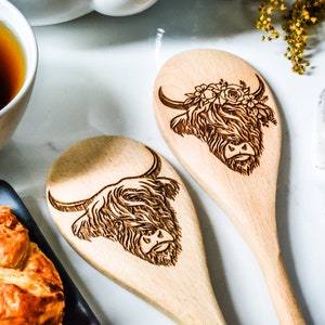 K&ouml;nnte beinhalten: Zwei Holzl&ouml;ffel mit gravierten Highland-Kuh-Designs. Eine Kuh hat einen Blumenkranz. Die L&ouml;ffel sind hellbraun und liegen auf einer wei&szlig;en Oberfl&auml;che, in der N&auml;he einer Tasse Tee und Geb&auml;ck. Ein charmantes K&uuml;chenutensil.