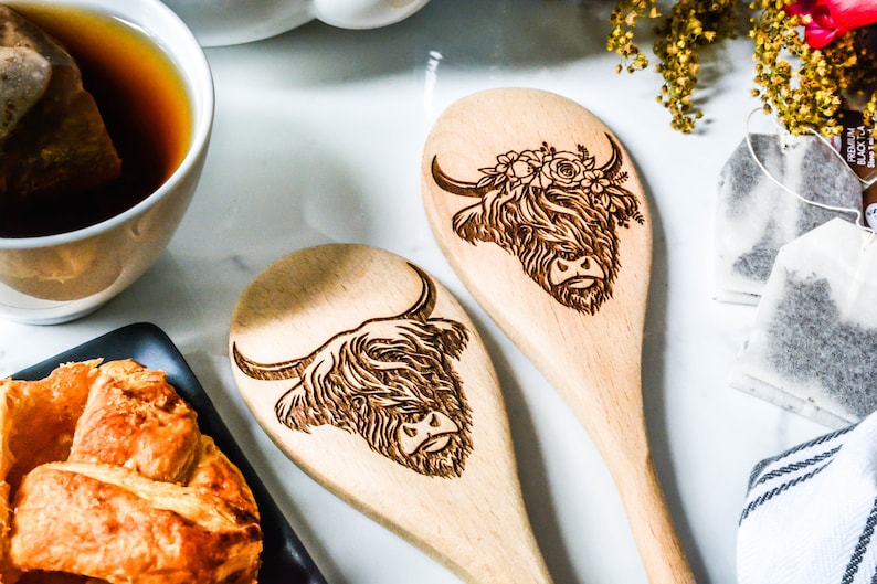K&ouml;nnte beinhalten: Zwei Holzl&ouml;ffel mit gravierten Highland-Kuh-Designs. Eine Kuh hat einen Blumenkranz. Die L&ouml;ffel sind hellbraun und liegen auf einer wei&szlig;en Oberfl&auml;che, in der N&auml;he einer Tasse Tee und Geb&auml;ck. Ein charmantes K&uuml;chenutensil.
