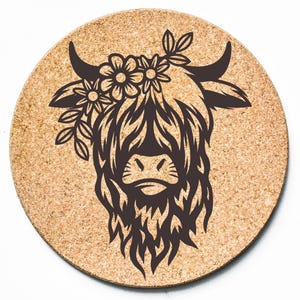 May include: Round cork coaster featuring a dark brown silhouette of a Highland cow's head. The cow has a floral crown and long, shaggy fur. The coaster is made of natural cork material.