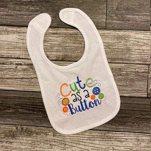 May include: White baby bib with the embroidered phrase "Cute as a Button" in orange, green, and blue. The bib features colorful button designs and a curved neckline.