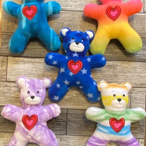 May include: A collection of colorful plush teddy bears. Each bear has a heart-shaped design on its chest. The bears are in various colors, including blue, purple, rainbow, and blue with white stars. The bears are soft and cuddly.