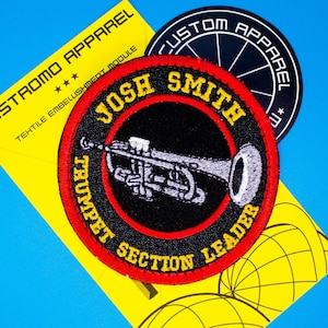 Pode incluir: Um remendo bordado em vermelho, branco e preto com um trompete e o texto "Josh Smith Trumpet Section Leader". O remendo está sobre um fundo amarelo com um design circular em preto e branco.