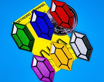 May include: A collection of embroidered patches in various jewel tones, including green, blue, red, yellow, purple, and gray. Each patch features a geometric gem design with white accents. A yellow card with text is visible.