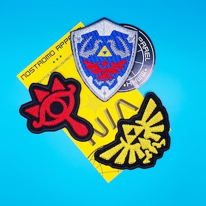 May include: Four embroidered patches featuring characters from the Legend of Zelda video game series. The patches are on a yellow background with the text "NOSTROMO APPAREL TEXTILE EMBELISHMENT" and "WWW.HHWWW.COM".