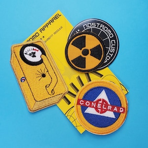 May include: Four embroidered patches, including a yellow patch with a black symbol and the text "Nostromo Customs", a yellow patch with a black symbol and the text "Conelrad", a yellow patch with a black symbol and the text "Assignment Module", and a yellow patch with a black symbol and the text "Nostromo Apparel".